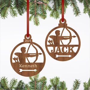 May include: Two wooden Christmas ornaments with red ribbons. Each ornament features a cutout design of an archer with a bow and arrow. One ornament has the name "Kenneth" and the other has "Jack". The year "2025" is on one ornament.