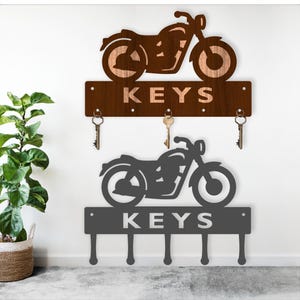Pode incluir: Dois porta-chaves com tema de motocicleta. O superior é castanho com a palavra "KEYS" recortada, e o inferior é cinzento com a palavra "KEYS". Ambos têm ganchos para chaves.