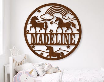 May include: A circular brown wooden wall sign featuring a cutout design of three unicorns, a rainbow, clouds, and stars. The name MADELINE is prominently displayed in capital letters across the center of the circular wooden frame.