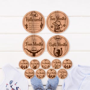 May include: A collection of wooden baby milestone discs with a cowboy theme. The discs are circular and display text like "Hello world," "Two Months," and "One Year." Each disc features a cowboy hat and other western-style graphics. The discs are arranged on a white surface.
