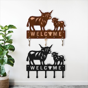 May include: Two key holders with Highland cow designs. One is brown, the other black. Both have the word "WELCOME" cut out. Each key holder has three hooks for keys, ideal for a hallway.