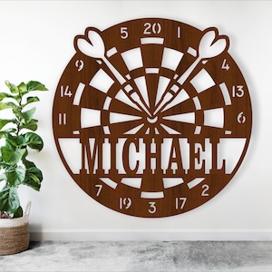 May include: A circular wooden dartboard in brown, with the name "MICHAEL" cut out in the centre. Two darts are shown on the board. Numbers 1 to 20 are around the edge. A green plant in a woven basket is to the left.