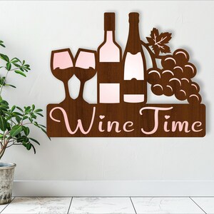 Könnte beinhalten: Wanddekoration aus Holz mit der Aufschrift "Wine Time" in rosa Buchstaben. Die Dekoration zeigt zwei Weingläser, zwei Weinflaschen und eine Traube.
