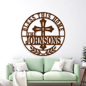 May include: A brown metal wall sign with the words "BLESS THIS HOME" in a circular design. The sign features a cross in the center and the family name "THE JOHNSONS" below. Decorative leaves are at the bottom of the sign.