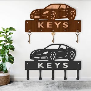 Puede incluir: Dos llaveros con forma de coche. El superior es marrón con la palabra "KEYS" recortada, y el inferior es negro con la palabra "KEYS" en blanco. Cada uno tiene ganchos para llaves.