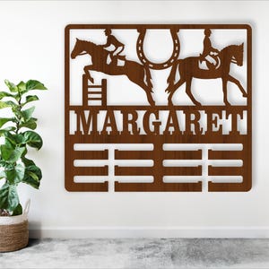 Op de afbeelding: Een bruine houten medailledisplay met de naam "MARGARET" erop gegraveerd. De display toont twee paarden en ruiters, één springt over een hek en de ander draaft. Er zijn acht vakken voor medailles.