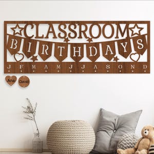 May include: A brown wooden classroom birthday chart with cutouts of stars, hearts, and banners that spell out "Classroom Birthdays". The chart has 12 slots for hanging name tags, with the months of the year labeled below. Two name tags are hanging on the chart, one that says "Ana" and one that says "David".
