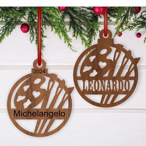 May include: Two wooden Christmas ornaments shaped like a circle with a paint palette and paintbrushes inside. The ornaments are personalized with the names "Michelangelo" and "Leonardo" and the year "2024".