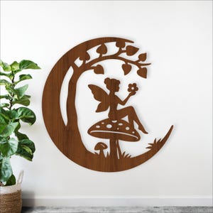 Op de afbeelding: Houten wandkunst met een fee die op een paddenstoel onder een boom zit, alles in een halve maanvorm. De fee houdt een bloem vast. Het kunstwerk heeft een warme bruine kleur en is op een witte muur bevestigd.