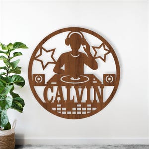 Puede incluir: Decoración de pared de madera que representa a un DJ con auriculares, estrellas y altavoces. El nombre "CALVIN" está recortado en el centro. La obra de arte es de color marrón cálido y está montada en una pared blanca. Ideal para amantes de la música.