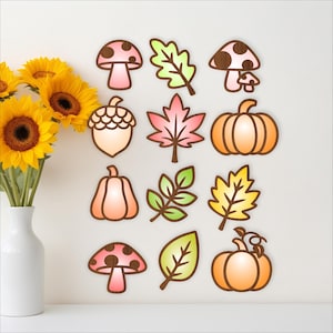 May include: A collection of fall-themed wooden cutouts, including pumpkins, leaves, mushrooms, and an acorn. The cutouts are in shades of orange, pink, green, and brown, with a white background. Sunflowers in a white vase are on the left.