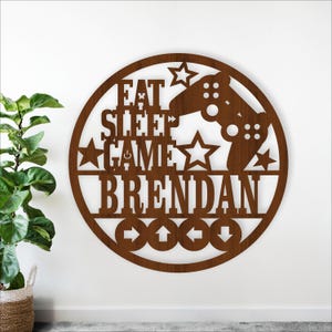 Op de afbeelding: Een bruin houten wandbord met de tekst "EAT SLEEP GAME BRENDAN" en een game controller ontwerp. Het bord is rond met ster- en pijluitsparingen. Een decoratief item voor een gamekamer.