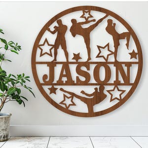 Puede incluir: Un letrero redondo de madera marrón con un diseño recortado de tres figuras en pose de lucha. El letrero tiene un fondo blanco y el nombre "JASON" está escrito en letras grandes en la parte inferior.