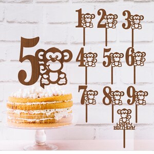 May include: Wooden cake toppers with monkey designs and numbers 1 through 9. The toppers are brown and have a white monkey face with black eyes. The topper for number 10 says "Happy Birthday".