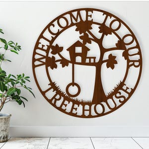 Könnte beinhalten: Ein braunes Holzschild mit den Worten "Willkommen in unserem Baumhaus" in einem Kreis um einen Baum mit einem Haus und einer Schaukel.