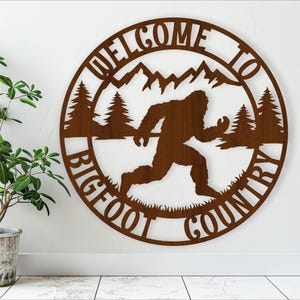 May include: A brown metal sign with the words "Welcome to Bigfoot Country" in a circle. The sign features a silhouette of Bigfoot walking through a forest.
