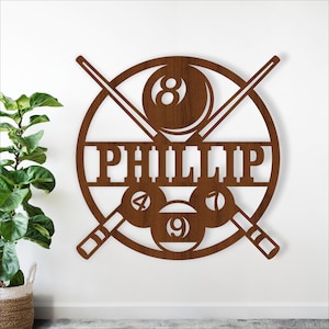 May include: A brown wooden wall art piece featuring a circular design with crossed pool cues. The design includes the name "PHILLIP" and pool balls numbered 4, 7, 8, and 9. The artwork is mounted on a white wall.