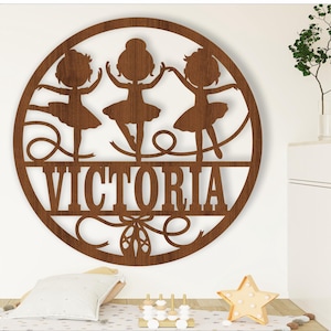 May include: A brown wooden round sign with three ballerina silhouettes and the name "VICTORIA" cut out. The sign is decorated with a bow and a pair of ballet shoes.