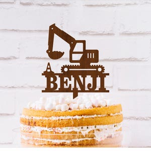 Könnte beinhalten: Brauner Holz-Kuchentopper mit einer Silhouette eines Baggers und dem Namen "Benji" darunter. Der Topper befindet sich auf einem mehrstöckigen Kuchen mit weißem Frosting und Marshmallows.