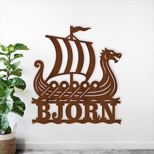 May include: A brown wooden Viking ship wall art with the name "BJORN" cut out at the bottom. The ship features a dragon head at the front, a sail, and oars. The art is mounted on a white wall.