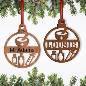 May include: Two wooden Christmas ornaments with a pharmacy theme. One ornament says "Mr. Adams" and the other says "LOUISE". Both ornaments feature a mortar and pestle, pills, and a syringe.
