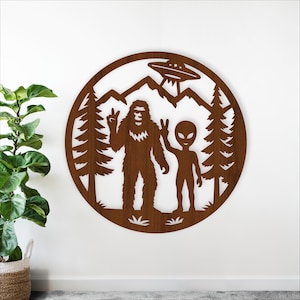 Op de afbeelding: Een ronde houten wanddecoratie met een Bigfoot en een alien die beide vredestekens tonen. Een UFO zweeft boven een bergketen, met dennenbomen aan weerszijden. Het kunstwerk is donkerbruin.