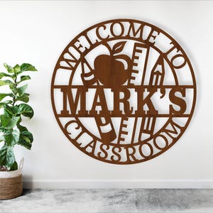 Könnte beinhalten: Ein rundes Schild aus braunem Holz mit dem Text "Willkommen in Marks Klassenzimmer" und einem Design aus Apfel, Bleistift und Lineal.