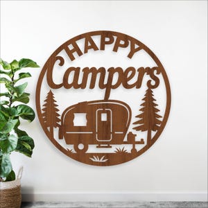 May include: A round, wooden sign with the words "HAPPY Campers" cut out. The sign features a camper, trees, and grass. The sign is brown and is likely for outdoor decor.
