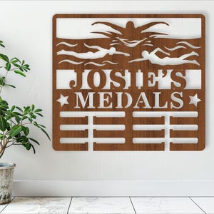 May include: A brown wooden medal display with a white silhouette of swimmers and the text "Josh's Medals" with stars on either side. The display has 10 slots for medals.