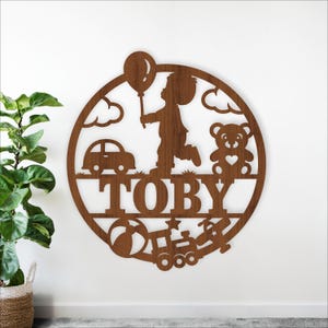 May include: A wooden wall decoration with the name "TOBY" cut out. The design includes a child with a balloon, a car, a teddy bear, clouds, a ball, a train, and a star, all within a circular frame, made of wood.