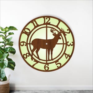 May include: A round wooden wall clock with a deer silhouette in the center. The clock face is light green, with brown numerals and a brown deer design. The clock has a classic, rustic style.