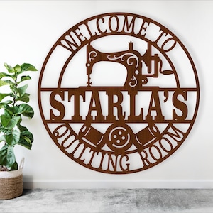 May include: A brown metal sign with the text "WELCOME TO STARLA'S QUILTING ROOM" in a circular design. The sign features a sewing machine, spools of thread, and a button. A potted plant is to the left.