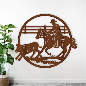 May include: A circular, brown metal wall art piece showing a cowboy on horseback herding a calf. The design includes a fence and grass, all within a circular frame. The artwork is suitable for rustic or Western-themed decor.