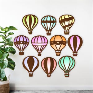 Op de afbeelding: Negen houten wanddecoraties in de vorm van luchtballonnen in verschillende kleuren, waaronder groen, blauw, roze en oranje. Elke ballon heeft een uniek ontwerp en is op een witte muur gemonteerd. De ballonnen zijn van hout en hebben een warme gloed.