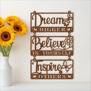 Peut inclure: Décoration murale en bois marron avec les mots « Dream Bigger », « Believe in Yourself » et « Inspire Others ». L'art présente des découpes de papillons et de fleurs. Un vase blanc avec des tournesols est au premier plan.