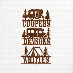 May include: A brown laser-cut wall sign featuring three stacked sections. Each section displays a camping vehicle—a travel trailer, a motorhome, and a tent—flanked by pine trees, with the names COOPERS, DENSONS, and WHITLES cut out below.