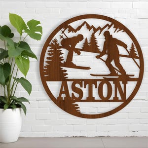 May include: A brown wooden round sign with a silhouette of two skiers on a mountain scene. The sign has the name "ASTON" written in large letters at the bottom.