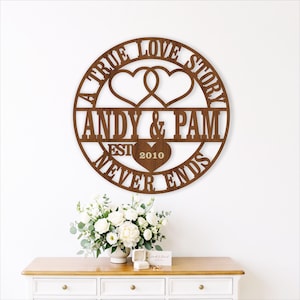 May include: A circular brown wooden wall sign featuring two interlocking heart outlines at the top, the names ANDY & PAM in the center, and the text A TRUE LOVE STORY NEVER ENDS surrounding the design with the year 2010 inside a heart.