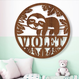 以下が含まれることがあります： 木製の円形ウォールデコレーション。 枝の上で眠るナマケモノのデザインで、葉で囲まれています。 下には「VIOLET」という名前が切り抜かれています。 茶色の木製部分は子供部屋に適しています。