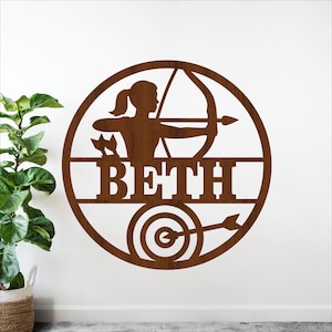 May include: A brown wooden wall art piece featuring a silhouette of a person aiming a bow and arrow. The name "BETH" is displayed below the archer, with a target and arrow at the bottom. The design is circular.