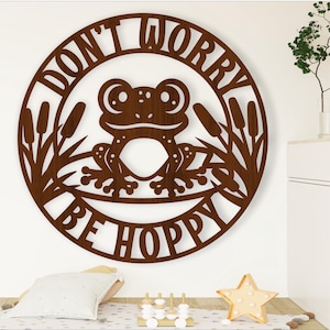 May include: Brown wooden wall decor with a circle design. The circle features a frog sitting on a lily pad with reeds around it. The text "Don't Worry Be Hoppy" is written around the frog.