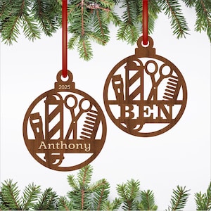 Peut inclure: Deux décorations de Noël en bois avec des motifs de barbier. Chaque ornement circulaire présente des outils de barbier découpés, les noms "Anthony" et "Ben", et l'année 2025. Ils sont suspendus à des rubans rouges sur un fond de branches de conifères.