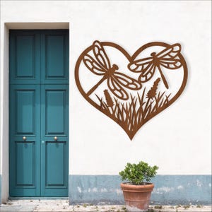 Puede incluir: Una pieza de arte mural de metal marrón con dos libélulas dentro de una forma de corazón. El diseño incluye detalles recortados de hierba y flores. La obra de arte está montada en una pared blanca cerca de una puerta turquesa y una planta en maceta.