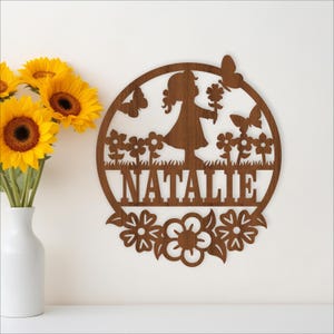 May include: A wooden wall decoration with a girl holding flowers, butterflies, and the name "NATALIE" cut out. The design is framed by flowers in a circular shape. A vase of sunflowers is to the left.