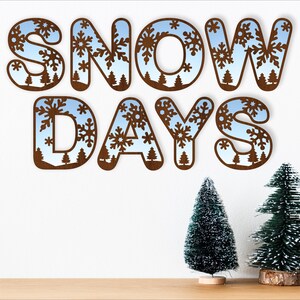 Peut inclure: Lettres en bois épellées "SNOW DAYS" avec un motif de flocons de neige bleus et blancs. Les lettres sont sur un mur blanc avec deux petits sapins de Noël verts devant elles.