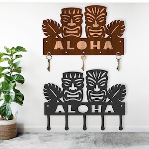 May include: Two metal key holders with tiki mask designs. The top one is brown with the word "ALOHA" and three key hooks. The bottom one is black with the word "ALOHA" and four key hooks. Decorative wall art.