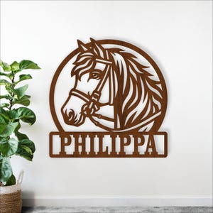 Könnte beinhalten: Wanddekoration aus Holz mit einem Pferdekopf im Profil, der einen Zaum trägt und in einem kreisförmigen Rahmen angeordnet ist. Der Name "PHILIPPA" wird darunter angezeigt. Die Kunst ist in einem warmen Braunton gehalten.