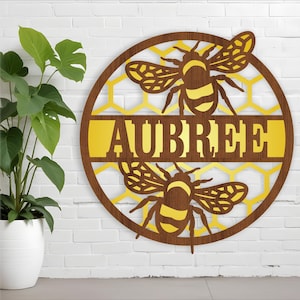 Könnte beinhalten: Ein rundes Holzschild mit einem Wabenmuster und zwei Bienen. Das Schild hat ein goldenes Band mit dem Namen "AUBREE" in braunen Buchstaben.