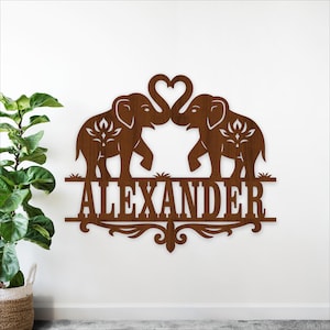 May include: A brown metal wall art piece featuring two elephants with trunks forming a heart shape. The name "ALEXANDER" is displayed below the elephants. Decorative floral elements are incorporated into the design.