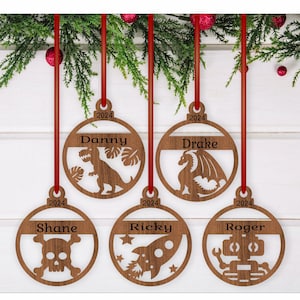 Könnte beinhalten: Fünf hölzerne Weihnachtskugeln mit Namen und Motiven. Die Kugeln sind rund und haben ein rotes Band befestigt. Die Namen auf den Kugeln sind Danny, Drake, Shane, Ricky und Roger. Die Motive auf den Kugeln sind ein Dinosaurier, ein Drache, ein Totenkopf mit Knochen, eine Rakete und ein Roboter.